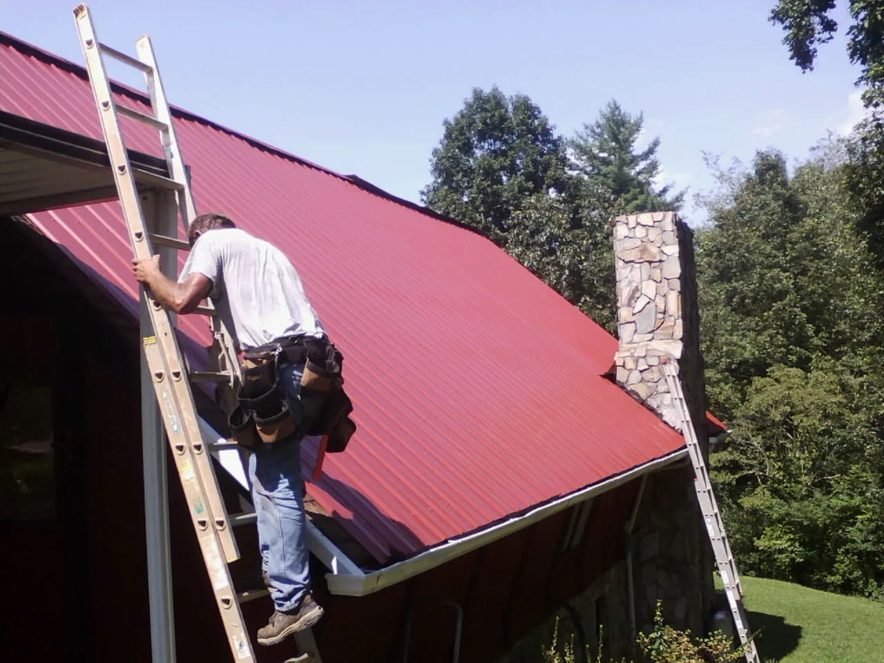 Roofer on a ladder beside a red metal roof and stone chimney — Soffit & Fascia Repair by Roofing Brothers in Freeland